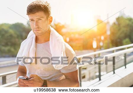 Stock Photo - Portrait of young attractive man standing with towel on his shoulders while using smartphone. Fotosearch