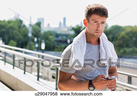 Stock Photo - Portrait of young attractive man standing with towel on his shoulders while using smartphone. Fotosearch