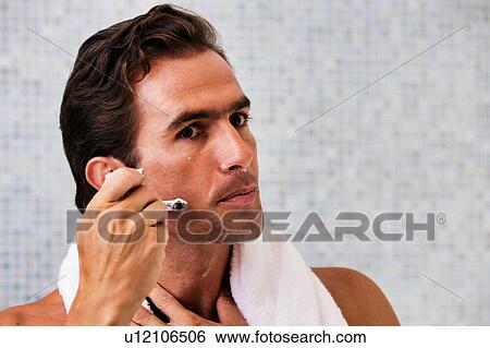 Stock Photograph - Portrait of young attractive man applying beard foam and shaving his beard while looking in the mirror. Fotosearch