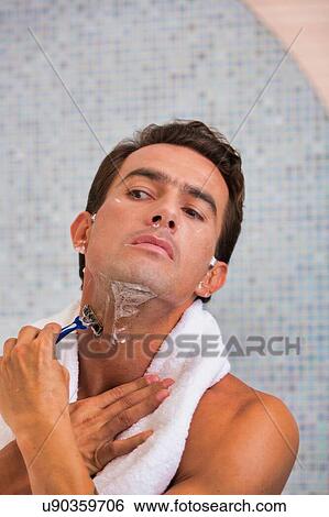 Stock Photograph - Portrait of young attractive man applying beard foam and shaving his beard while looking in the mirror. Fotosearch