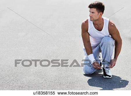 Stock Photography - Portrait of young attractive man wearing sports wear tying his rubber shoes in park. Fotosearch