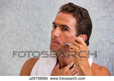 Stock Photography - Portrait of young attractive man applying beard foam and shaving his beard while looking in the mirror. Fotosearch