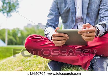Picture - Portrait of young attractive businessman using digital tablet while sitting on the grass. Fotosearch