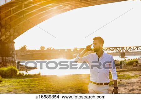 Picture - Portrait of young attractive businessman drinking beer and walking barefoot on the beach while holding his shoes. Fotosearch