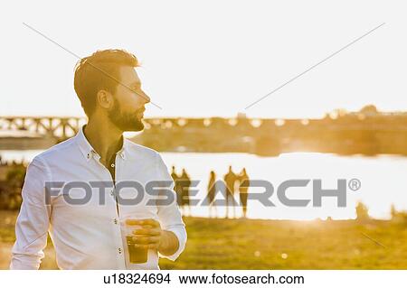 Picture - Portrait of young attractive businessman drinking beer and walking barefoot on the beach while holding his shoes. Fotosearch