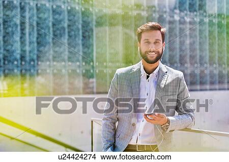 Picture - Portrait of young attractive businessman using smartphone with earphones on. Fotosearch
