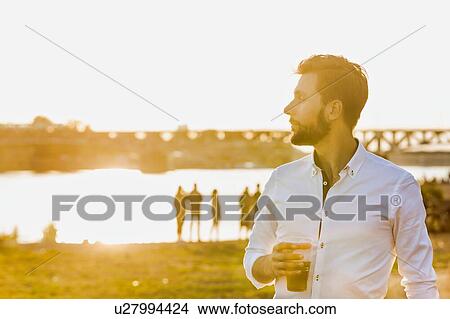 Picture - Portrait of young attractive businessman drinking beer and walking barefoot on the beach while holding his shoes. Fotosearch