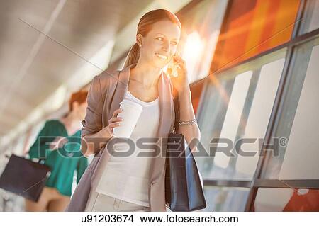 Picture - Portrait of young attractive businesswoman talking on smartphone and holding cup of coffee while walking against colleague. Fotosearch