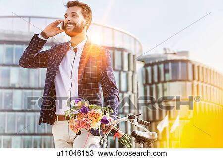 Stock Image - Portrait of young attractive businessman holding boquet of flowers while talking on smartphone. Fotosearch
