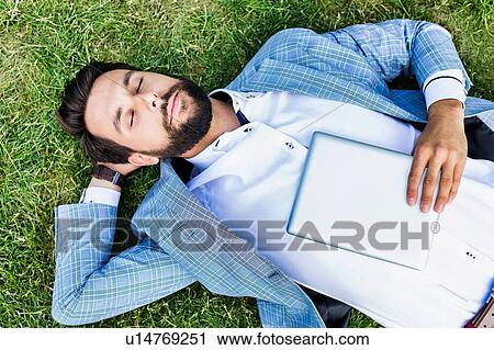 Stock Image - Portrait of young attractive businessman holding digital tablet while sleeping and lying on the grass. Fotosearch