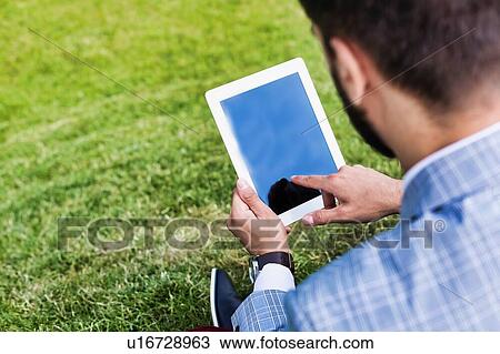 Stock Image - Portrait of young attractive businessman using digital tablet while sitting on the grass. Fotosearch