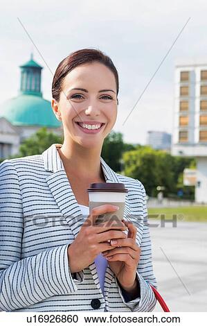 Stock Image - Portrait of young attractive businesswoman smiling while holding a cup of coffee outside office building. Fotosearch