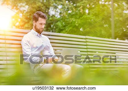 Stock Image - Portrait of young attractive businessman sitting on bench while working on his laptop. Fotosearch