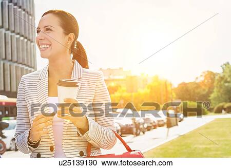 Stock Image - Portrait of young attractive businesswoman smiling while holding a cup of coffee outside office building. Fotosearch