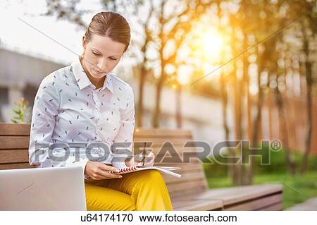 Stock Image - Portrait of young attractive businesswoman writing note and looking on laptop while sitting on bench. Fotosearch