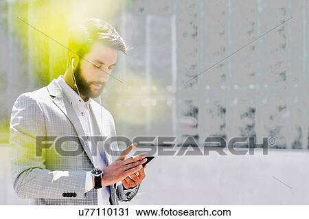 Stock Image - Portrait of young attractive businessman using smartphone with earphones on. Fotosearch