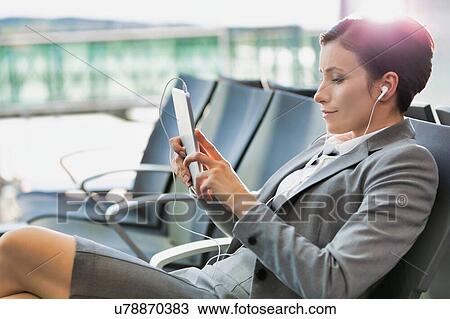 Stock Image - Portrait of young attractive businesswoman using digital tablet with earphones on while waiting for boarding in airport with lens flare. Fotosearch
