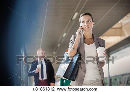 Stock Image - Portrait of young attractive businesswoman talking on smartphone and holding cup of coffee while walking against colleague. Fotosearch