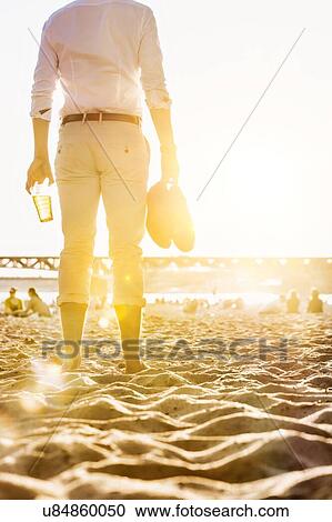 Stock Image - Portrait of young attractive businessman drinking beer and walking barefoot on the beach while holding his shoes. Fotosearch
