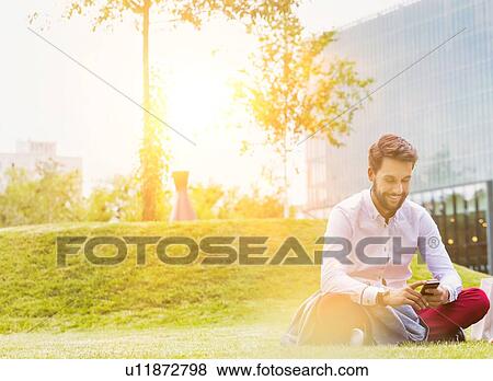 Portrait of young attractive businessman using smartphone while sitting on the grass View Large Photo Image Stock Photo - Portrait of young attractive businessman using smartphone while sitting on the grass. Fotosearch