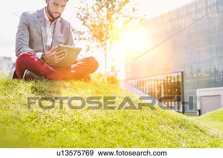 Stock Photo - Portrait of young attractive businessman using digital tablet while sitting on the grass. Fotosearch