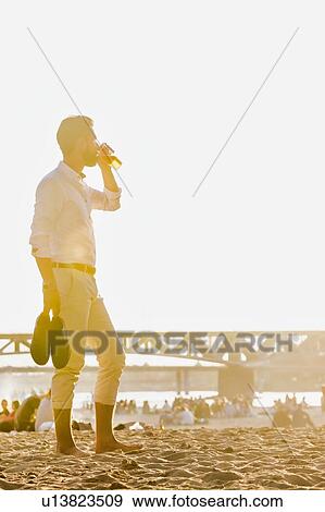 Stock Photo - Portrait of young attractive businessman drinking beer and walking barefoot on the beach while holding his shoes. Fotosearch