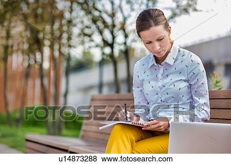 Portrait of young attractive businesswoman writing note and looking on laptop while sitting on bench View Large Photo Image Stock Photo - Portrait of young attractive businesswoman writing note and looking on laptop while sitting on bench. Fotosearch