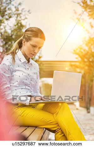 Portrait of young attractive businesswoman sitting while working on her laptop and sitting on bench View Large Photo Image Stock Photo - Portrait of young attractive businesswoman sitting while working on her laptop and sitting on bench. Fotosearch