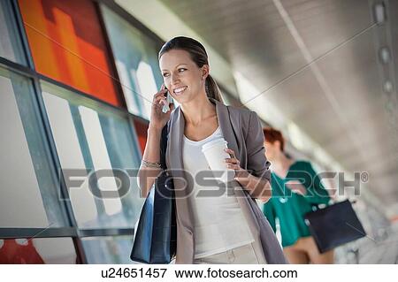 Stock Photo - Portrait of young attractive businesswoman talking on smartphone and holding cup of coffee while walking against colleague. Fotosearch