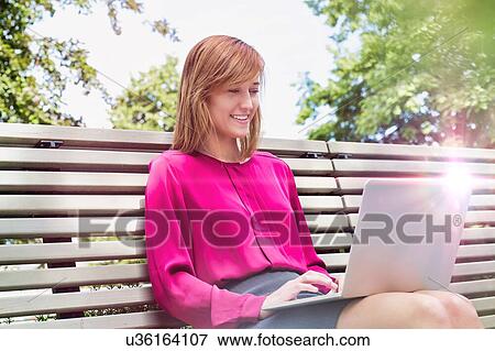 Portrait of young attractive businesswoman sitting on bench while working on her laptop View Large Photo Image Stock Photo - Portrait of young attractive businesswoman sitting on bench while working on her laptop. Fotosearch