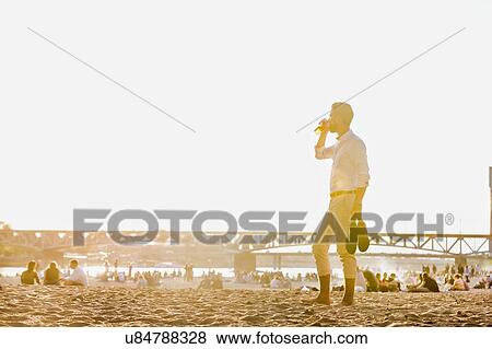 Stock Photo - Portrait of young attractive businessman drinking beer and walking barefoot on the beach while holding his shoes. Fotosearch
