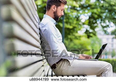 Stock Photo - Portrait of young attractive businessman sitting on bench while working on his laptop. Fotosearch