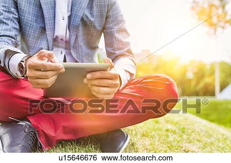 Stock Photograph - Portrait of young attractive businessman using digital tablet while sitting on the grass. Fotosearch
