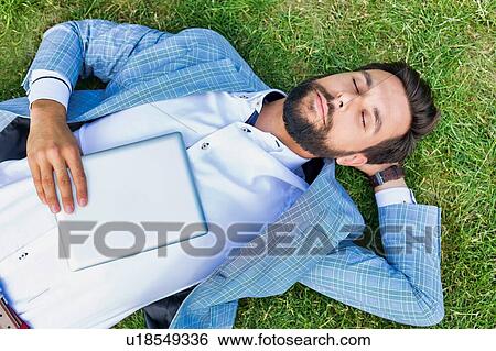 Stock Photograph - Portrait of young attractive businessman holding digital tablet while sleeping and lying on the grass. Fotosearch