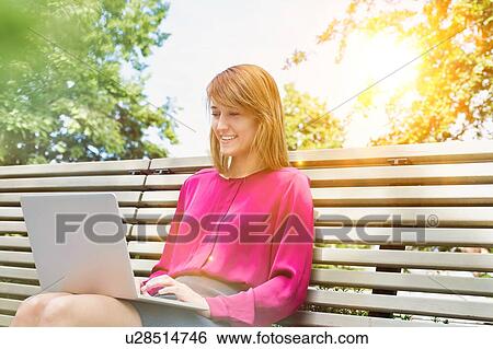 Stock Photograph - Portrait of young attractive businesswoman sitting on bench while working on her laptop. Fotosearch