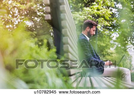 Portrait of young attractive businessman sitting on bench while working on his laptop View Large Photo Image Stock Photography - Portrait of young attractive businessman sitting on bench while working on his laptop. Fotosearch