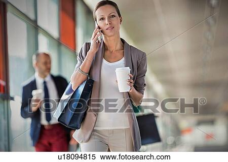 Stock Photography - Portrait of young attractive businesswoman talking on smartphone and holding cup of coffee while walking against colleague. Fotosearch