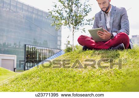 Stock Photography - Portrait of young attractive businessman using digital tablet while sitting on the grass. Fotosearch
