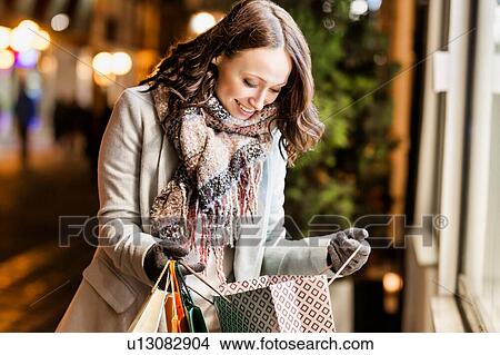 Picture - Portrait of young attractive woman smiling while looking at her new clothes on paper bag . Fotosearch