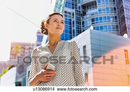 Picture - Portrait of young attractive woman holding smartphone while standing against office building with lens flare . Fotosearch