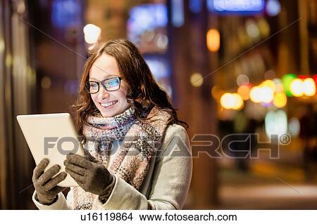 Picture - Portrait of young attractive woman smiling and using digital tablet while on night shopping. Fotosearch