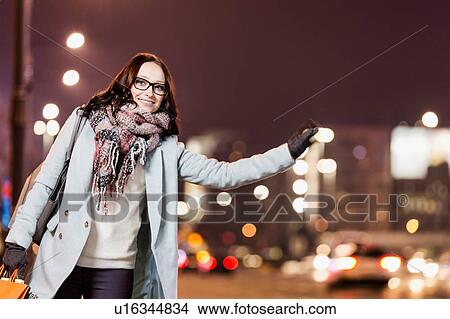 Picture - Portrait of young attractive woman standing while calling for taxi after night shopping. Fotosearch