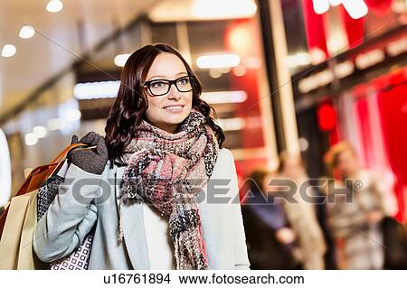 Picture - Portrait of young attractive woman walking while holding paper bags on night shopping. Fotosearch