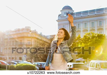 Stock Image - Portrait of young attractive woman calling taxi in the middle of the city with lens flare in background. Fotosearch