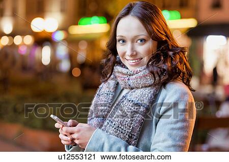 Stock Image - Portrait of young attractive woman smiling while using smartphone. Fotosearch