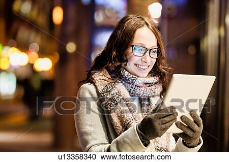 Stock Image - Portrait of young attractive woman smiling and using digital tablet while on night shopping. Fotosearch