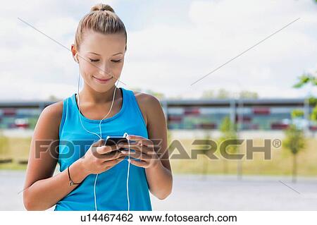Stock Image - Portrait of young attractive woman standing while using and listening to music on her smartphone in park. Fotosearch