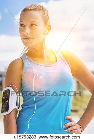 Stock Image - Portrait of young attractive woman standing while listening to music on her smartphone in park. Fotosearch