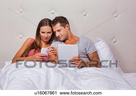 Stock Image - Portrait of young attractive woman showing smartphone while his boyfriend playing games on his digital tablet in bed. Fotosearch