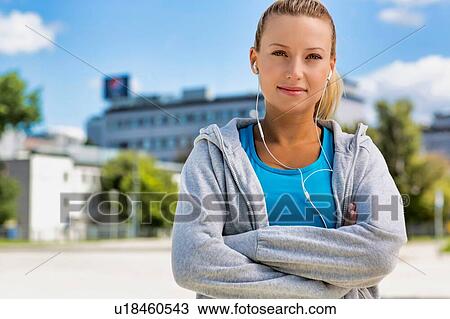 Stock Image - Portrait of young attractive woman standing with arms crossed while listening to music on her smartphone in park. Fotosearch